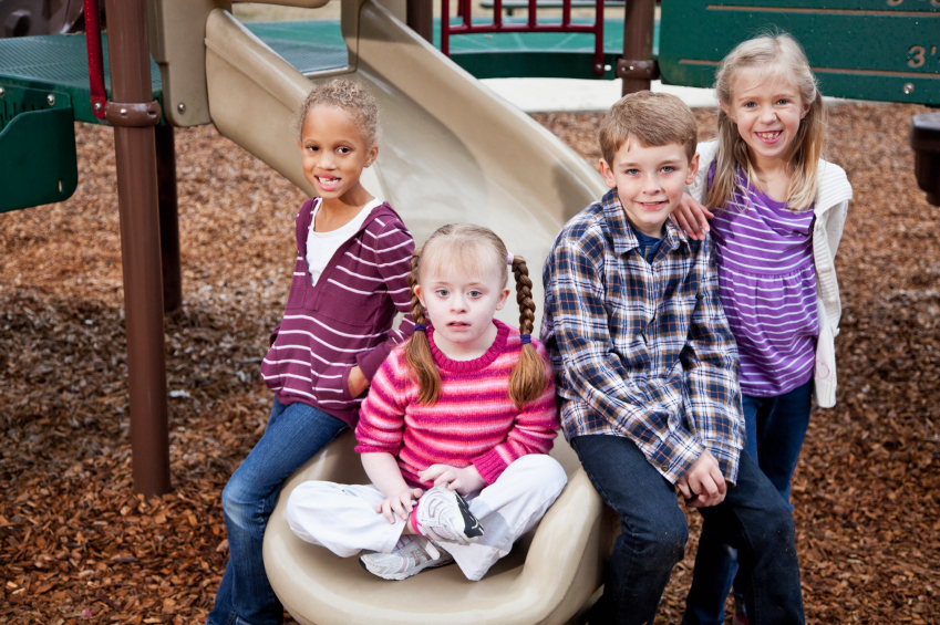 Multi-ethnic group of children on playground, ages 7 to 9. Girl sitting on slide has down syndrome.